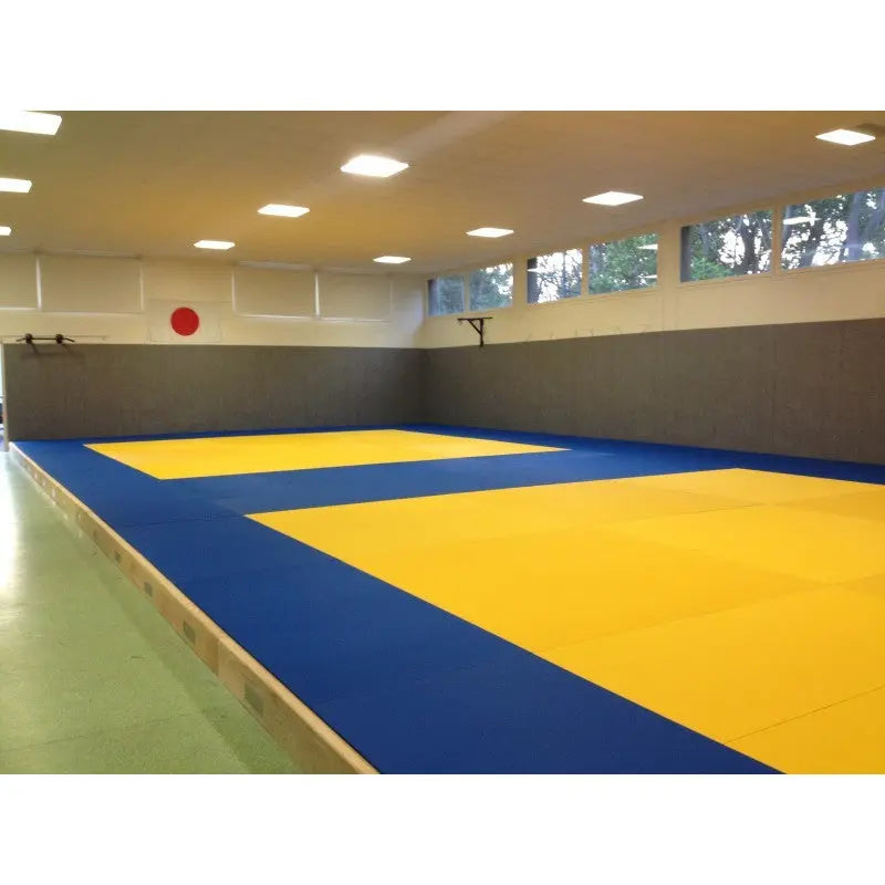 SportCom Judo-Tatami Reisstroh 1 × 1 m, 5 cm, rutschfeste Unterseite, blaue Fläche mit gelbem Feld in einer Halle mit Fensterband