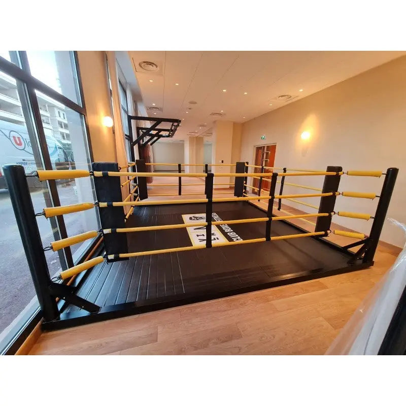 SportCom Freistehender Boxring mit gelben Seilpolstern, Frontansicht in einem Raum mit Holzboden und großen Fenstern