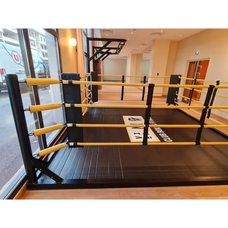 SportCom Freistehender Boxring mit gelben Seilen, Seitenansicht entlang der Fensterfront eines hellen Trainingsraums