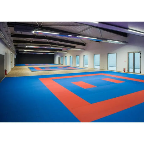 SportCom Judo-Tatami Reisstroh 1 × 1 m, 5 cm, rutschfeste Unterseite, blaue Fläche mit rotem Quadrat in einer langen Halle