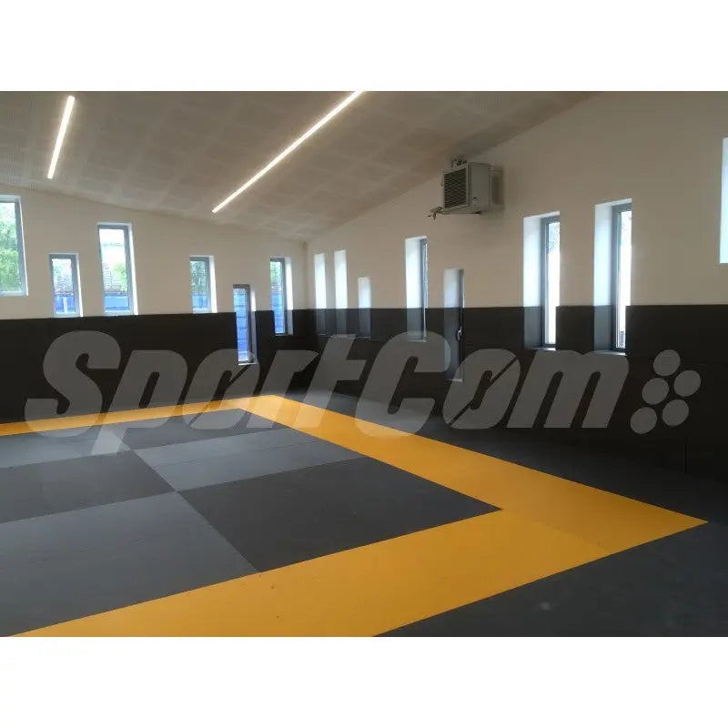 SportCom Judo-Tatami Reisstroh 1 × 1 m, 5 cm, rutschfeste Unterseite, schwarze Fläche mit gelbem Rahmen in einer Halle
