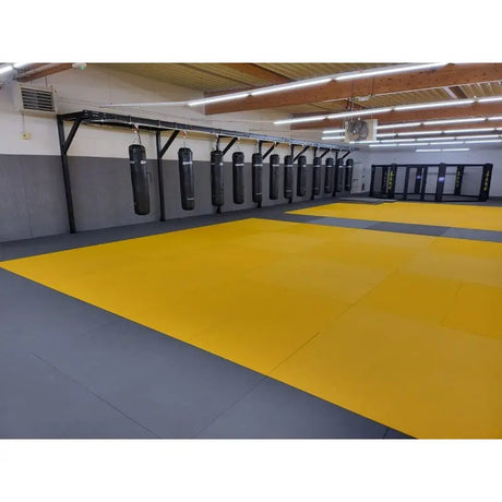 SportCom Judo-Tatami Reisstroh 2 × 1 m, 5 cm, glatte Unterseite, gelbe Fläche mit grauem Rand in Halle mit Sandsäcken